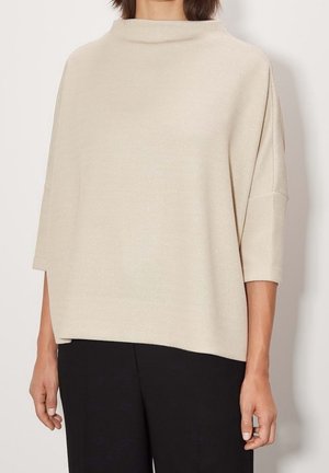Femme portant un pull beige ample à manches longues avec un col montant et un pantalon noir, debout devant un fond uni.
