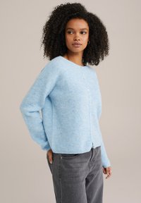 Ljust blå cardigan i mjukt, texturerat stickat tyg, med knappstängning och ribbad detalj på ärmar och nederkant. Avslappnad passform.
