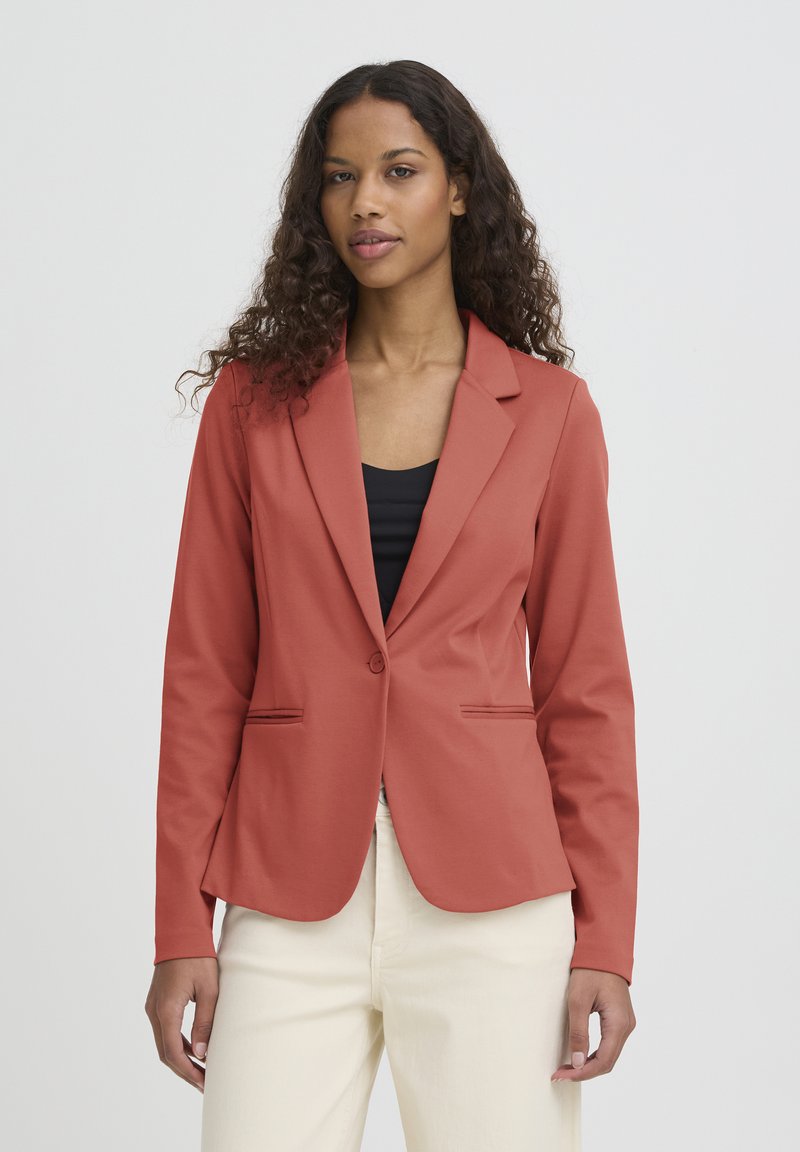 Veste couleur rouille avec une coupe ajustée, des revers crantés, deux poches devant et une fermeture à un bouton, associée à un haut noir.