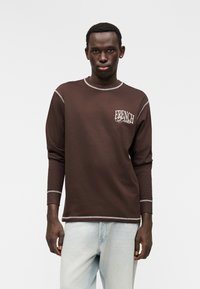 Καφέ μακρυμάνικο πουκάμισο με λευκές ραφές και το γράφημα "FRENCH" στο στήθος, σε συνδυασμό με ανοιχτόχρωμο τζιν παντελόνι, μοντέλο που στέκεται μπροστά σε λευκό φόντο.