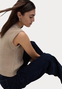 Beige ärmlös tröja med kabelstickad design och ribbad kant, i kombination med mörkblå vida byxor, sittande position mot en enfärgad bakgrund.