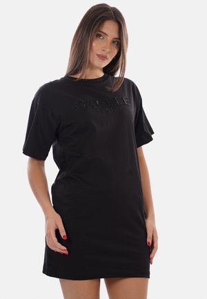 Femme aux cheveux bruns et lisses portant une robe t-shirt noire oversize avec le texte « GAELLE » sur la poitrine, debout devant un fond uni.