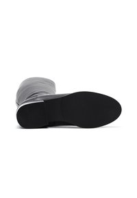 Zwarte leren kniehoog laars die plat ligt met de zool naar boven, waarbij de gestructureerde hak en de gladde buitenzool zichtbaar zijn op een witte achtergrond.