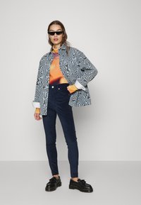 Casaco cinza com padrão, camiseta gráfica laranja e roxa por baixo, jeans escuros texturizados e justos, e sapatos de plataforma pretos.