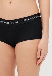 Svarte ribbede boyshorts med bred elastisk midje som har teksten "COPENHAGEN STUDIOS" i hvitt. Mykt stoff, tettsittende design.