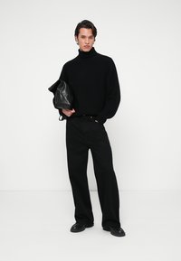 Un pull à col roulé noir à côtes assorti à un pantalon noir ample. L'individu tient un sac noir texturé, avec une finition lisse et des détails subtils.