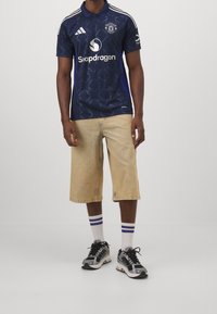 Granatowa koszulka piłkarska z geometrycznym wzorem, trzy białe paski na ramionach, zestawiona z beżowymi szortami do kolan i czarnymi trampkami.