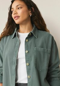Oversized groene denimbloes met een voorzak, knoopsluiting en een kraag, gelaagd over een witte ribbeltop.