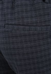 Tmavě šedé kalhoty s texturovaným vzorem, vybavené jednou kapsou a elastickým pasem pro pohodlí. Materiál vypadá měkce a flexibilně.