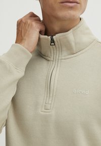 Sudadera beige con cremallera y cuello alto, puños acanalados y un pequeño logo. Tela suave con un brillo sutil y costuras detalladas.