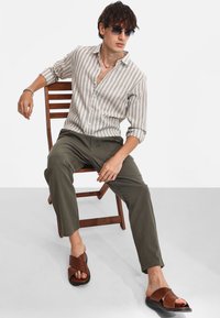 Camicia a righe grigio chiaro e bianca con bottoni, pantaloni oliva e sandali marroni con bretelle incrociate. Gli occhiali da sole e una sedia di legno completano il look.