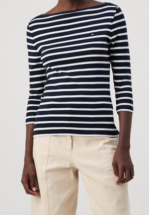 Marineblauwe en witte gestreepte longsleeve met een bootnek en een klein logoaccent. Gecombineerd met lichtbeige broeken. Zacht, rekbaar materiaal.