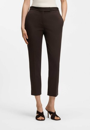 Frau trägt dunkelbraune, kurz geschnittene Hose mit Fronttaschen und schwarze Sandaletten mit hohen Absätzen, stehend vor weißem Hintergrund.