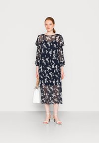 Vestido floral azul marinho com mangas longas transparentes e um design em camadas, apresentando pequenas flores brancas e uma silhueta fluida. Bolsa branca e saltos altos.