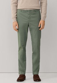 Groene katoenen broek, slim fit, met nette voorzakken, ritssluiting en riemlussen, gecombineerd met bruine suède schoenen.