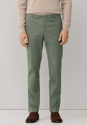 Groene katoenen broek, slim fit, met nette voorzakken, ritssluiting en riemlussen, gecombineerd met bruine suède schoenen.