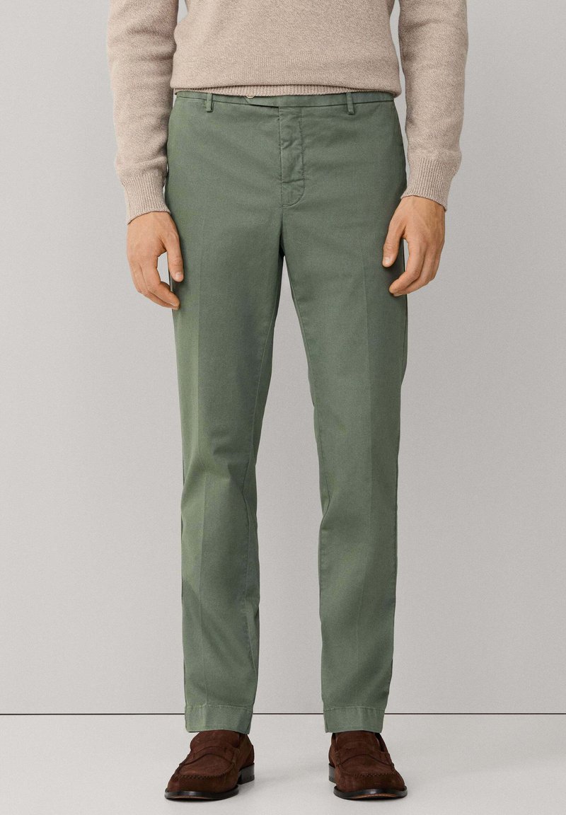 Groene katoenen broek, slim fit, met nette voorzakken, ritssluiting en riemlussen, gecombineerd met bruine suède schoenen.