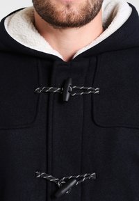 Uomo che indossa un cappotto nero con foderatura in pile bianca e due chiusure a toggles neri con cordini bianco e nero.