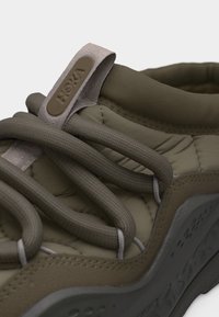 Κοντινό πλάνο ελιάς πράσινου αθλητικού παπουτσιού με χοντρές στρογγυλές κορδόνια, επενδεδυμένο ύφασμα στην επάνω πλευρά και ταμπελάκι με τη μάρκα στη γλώσσα.