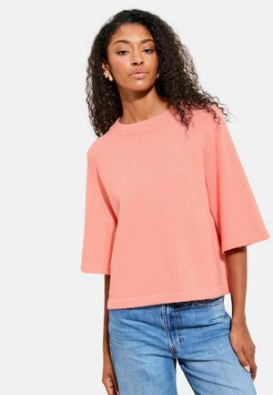 Jeune femme aux cheveux bouclés portant des boucles d'oreilles en or, un pull ample à manches courtes corail rose, et un jean bleu clair sur un fond blanc.