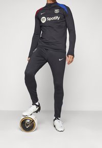 Schwarzer Nike Trainingsanzug mit einem Oberteil mit Viertelreißverschluss, das blaue und bordeauxrote Akzente, das Barcelona-Logo und das Spotify-Logo aufweist. Modell steht auf einem Fußball.