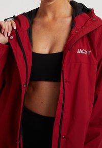 Femme portant une veste rouge ouverte avec une capuche et un soutien-gorge de sport noir, montrant le ventre, tenant le col de la veste d'une main.