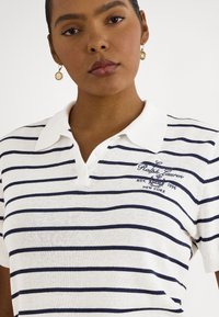 Sieviete tērpusies baltā polo krekliņā ar tumši ziliem horizontāliem svītrām un izšūtu uzrakstu "Lauren Ralph New York", nēsā zelta apaļas auskarus.