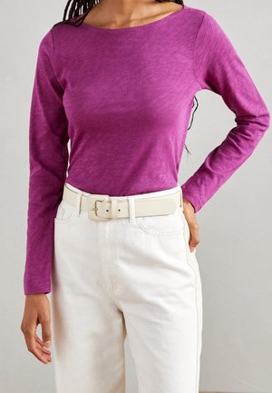 Kobieta ubrana w dopasowaną, długorękawową, fioletową bluzkę włożoną w białe spodnie z wysokim stanem i szerokim białym paskiem.