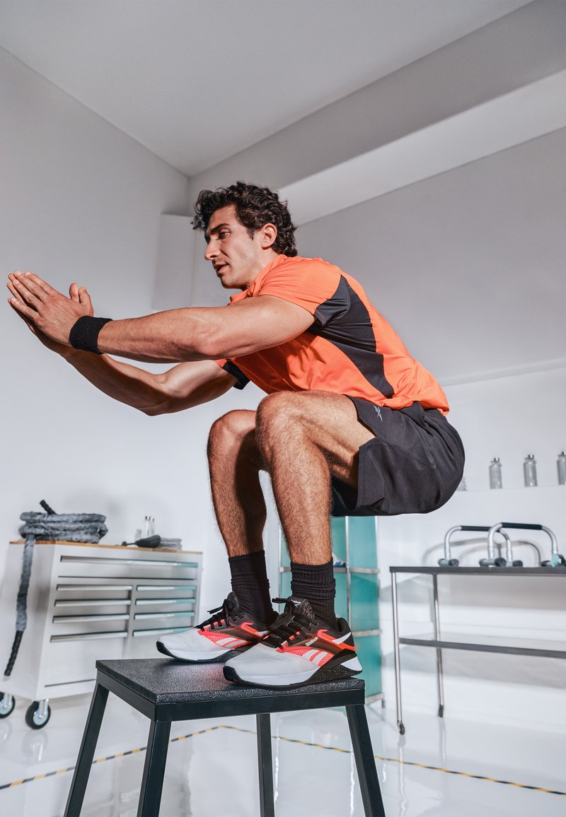 Un atleta masculino realiza una sentadilla en un taburete negro, vistiendo una camiseta naranja brillante y negra, pantalones cortos negros, y zapatillas deportivas negras y rojas.