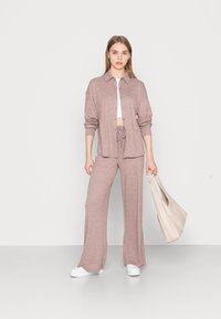 Conjunto de malha rosa claro, composto por uma camisa com gola e botões e calças com cordão combinando. A modelo segura uma mala bege e usa sapatos brancos.