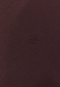 Burgundy sweatshirt med ett texturerat stickat mönster och en liten broderad logotyp i matchande färg på bröstet. Mjuk, avslappnad tyg.