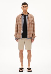 Chemise à carreaux marron surdimensionnée superposée à une chemise boutonnée grise, un t-shirt noir, un short beige et des sandales noires. Look simple et décontracté.