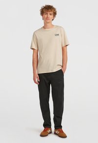 T-shirt in cotone beige con maniche corte e scollo rotondo, caratterizzato da un piccolo logo nero. Abbinato a pantaloni neri e scarpe in suede marroni.