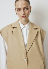 Beige sleeveless blazer dengan kerah berbentuk notch, penutupan satu kancing, dan saku samping, dilapisi di atas kemeja putih berkerah. Tekstur halus.