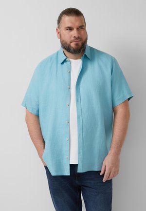 Brodaty mężczyzna w jasno-niebieskiej koszuli z krótkim rękawem na białej koszulce, stojący ze włożonymi w kieszenie rękami na jednolitym tle.