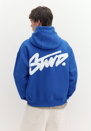 Jeune homme de dos portant un sweat à capuche bleu vif avec un grand texte blanc stylisé dans le dos et un pantalon beige clair.