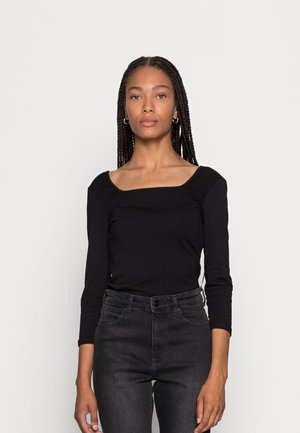 Schwarzes Langarmoberteil mit quadratischem Ausschnitt und figurnaher Passform, aus weichem Stoff, kombiniert mit hoch sitzenden dunklen Jeans.