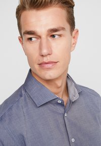 Koszula zapinana na guziki w niebieskim materiale chambray, z wzorzystym białym i granatowym kołnierzykiem w paski. Dwa guziki widoczne na szyi.