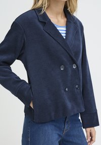 Veste en velours côtelé bleu marine à double boutonnage, col cranté et gros boutons. Présente une coupe carrée et une texture côtelée.