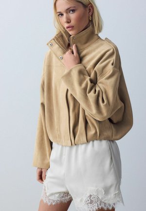 Naine kannab põldpunases seemisnahast tõmblukuga jopet ja valge pitsidega ääristatud lühikseid pükse, hoides ühte kätt jope krae peal.