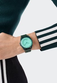 Grön silikonklocka med en mintgrön urtavla, cirkulär skiva, tim- och minutvisare samt ett mönstrat armband, bärs på ett handled.