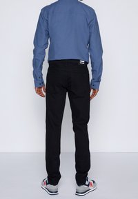 Blaues Langarmhemd mit Knopfmanschetten, kombiniert mit schwarzen Jeans und grauen Turnschuhen. Die Jeans haben ein Etikett am Bund und zwei Gesäßtaschen.