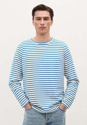 Tricou cu maneci lungi, în dungi albastre și albe, cu decolteu rotund și logo brodat la margine, asortat cu blugi albaștri deschisi.