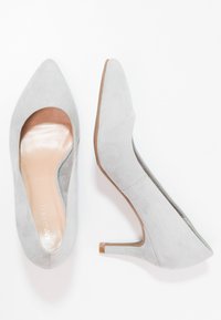 Šedé semišové lodičky na vysokém podpatku s špičatou špičkou a hladkou, světlou vnitřní stélkou. Mají výřezový design a štíhlý dřevěný podpatek.