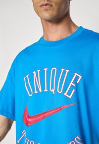 Mėlynas medvilnės marškinėliai su apvalia iškirpte, ant priekio yra ryškus baltas tekstas ir raudonas Nike logotipas. Trumpi rankovės, laisvas siluetas.