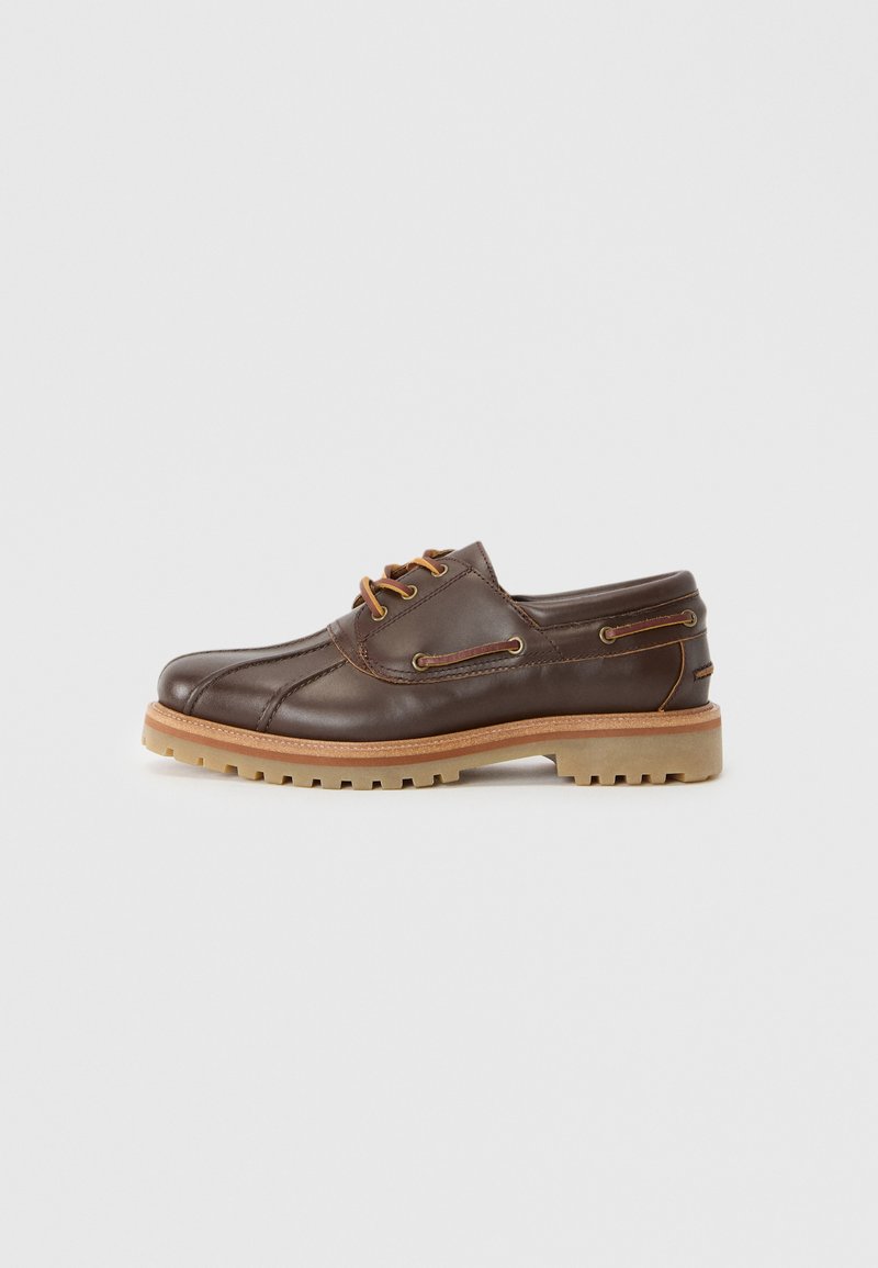 Chaussure à lacets en cuir marron avec une finition texturée, semelle en caoutchouc de couleur crème, coutures décoratives et accents en métal sur les côtés.