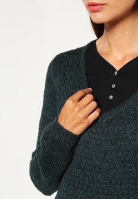 Dark green knitted sweater with a V-neck design, texture featuring a pattern, worn over a black button-up shirt with round buttons.