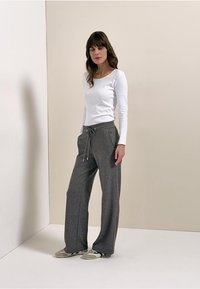 Weißes Langarmshirt und graue, weit geschnittene Jogginghosen mit Kordelzug. Das Model steht vor einem neutralen Hintergrund, die Schuhe sind grau und grün.