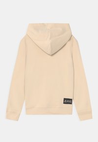 Sudadera con capucha beige que presenta una capucha con cordón, puños y borde acanalados, con una etiqueta cuadrada negra con la palabra "JEANS" en el lado inferior izquierdo.