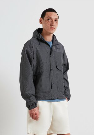 Junger Mann mit Sommersprossen, der eine schwarze karierte Kapuzenjacke, ein blaues Hemd und weiße Shorts trägt und vor einem schlichten Hintergrund steht.